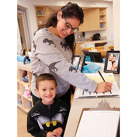 Preschool classroom, mother writing in parent sign-in binder, her son next to her