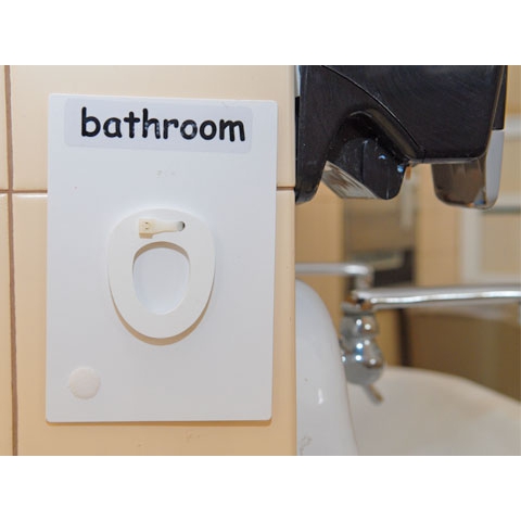 Sign that reads Bathroom, with physical miniature toilet seat and written text on wall near bathroom